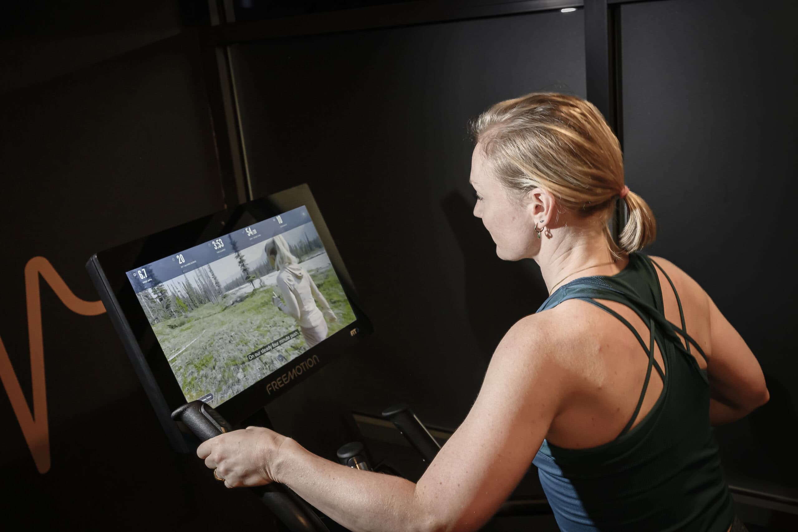 A woman using a stationary exercise bike with an interactive screen displaying a scenic outdoor trail featuring a runner. She is wearing a teal workout top with strappy back detailing, focused on her workout in a dimly lit gym environment.