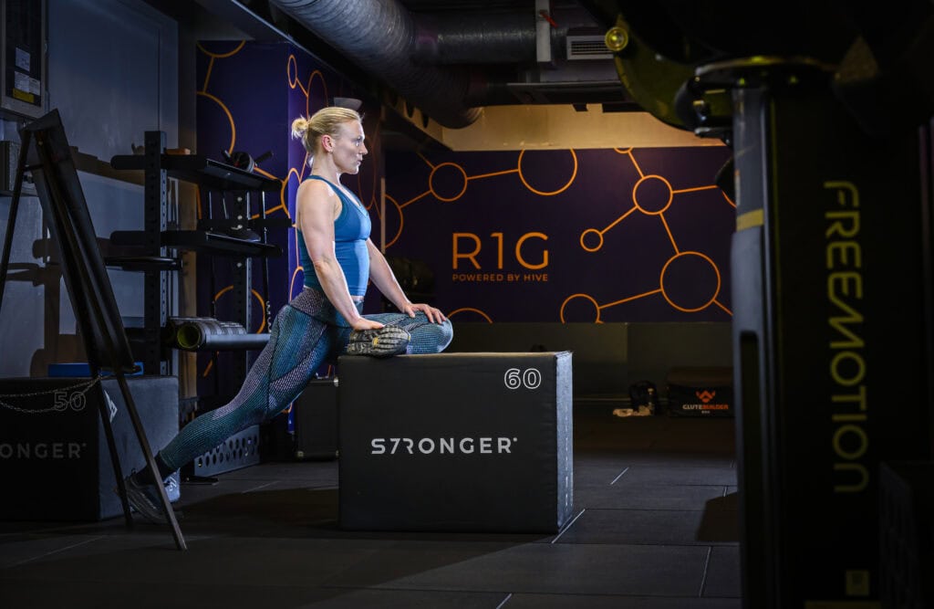 A woman stretching her leg on a sturdy workout box labeled 'STRONGER 60' in a modern gym environment. She is wearing a blue tank top and patterned leggings, with the gym branding 'R1G Powered by Hive' visible in the background. The space is equipped with gym equipment and features a dark, industrial design with vibrant orange accents.