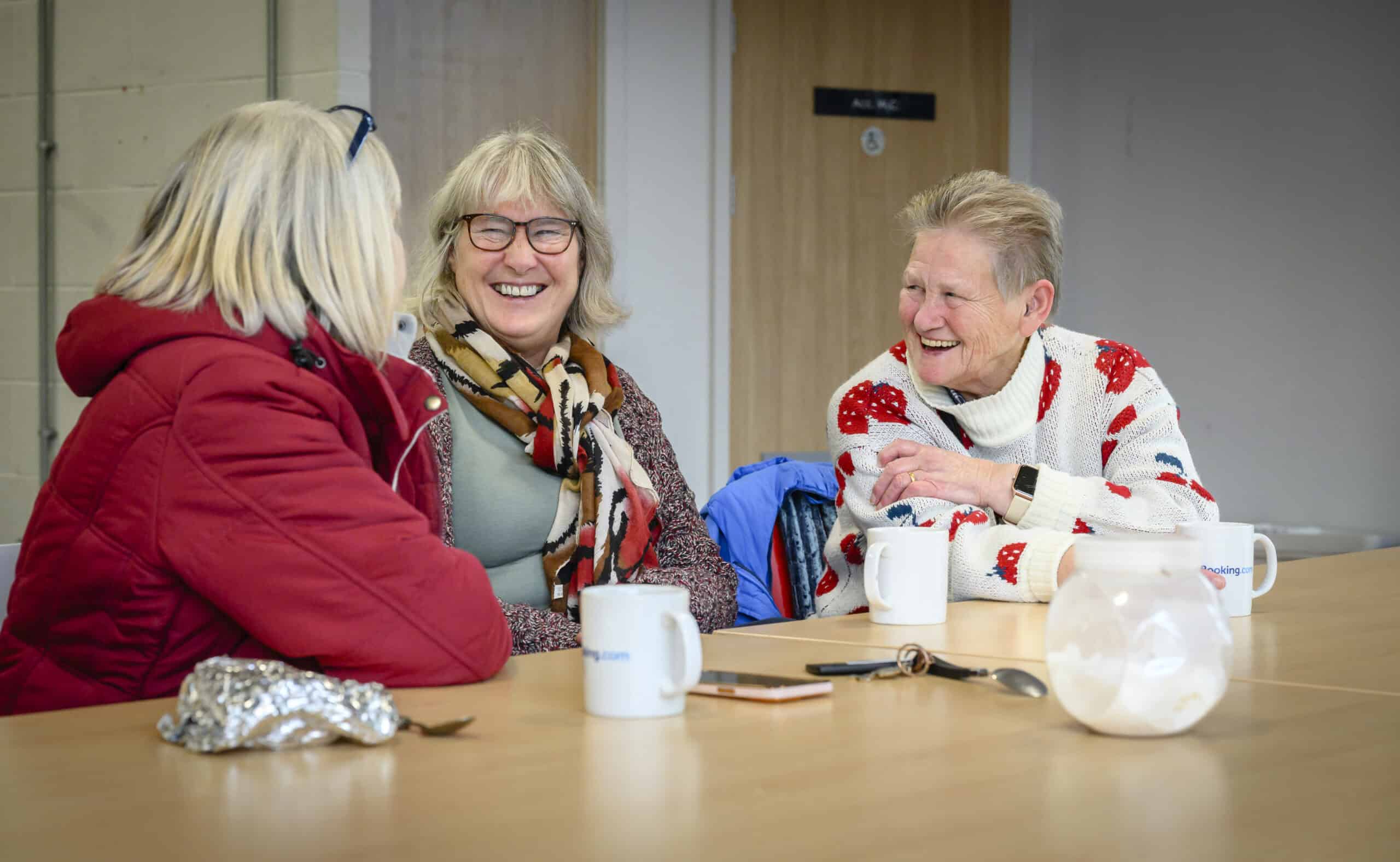 Three older women sitting at a table, smiling and chatting over cups of tea. One woman is wearing a red coat, another a patterned scarf, and the third a white sweater with red and blue designs. A phone, a spoon, and a sugar jar are on the table in front of them.